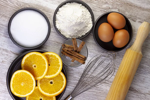 INGREDIENTS AND KITCHEN UTENSILS FOR PASTRY. ORANGE, FLOUR, SUGAR, EGGS AND CINNAMON NEXT TO WOOD AND ROLLER WOOD