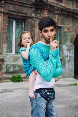 Komik baba ve kız parmaklarıyla şşşş yapar, dudaklarını yakın tutar, şehrin arka planında mavi kumaş taşıyıcı eşarp kullanır.