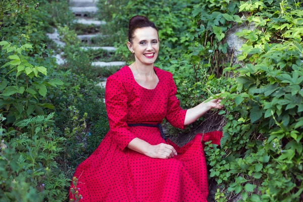 Portrait of smiling woman 50 years old wearing elegant red dress with dots, charming retro vintage look, with  green bushes around, sit on stairs