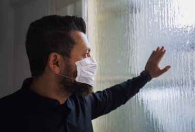 Koruyucu tıbbi maskeli bir adam pencereye dokunur, karantinada kalır, ciddi bir yüz, dünya koronası virüsü