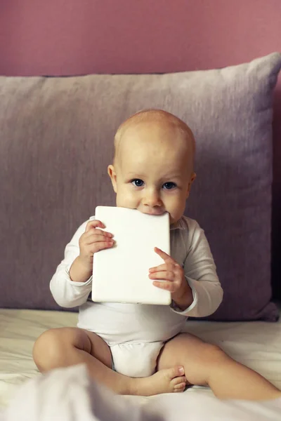 可爱的婴儿抱着和咬着电子书阅读器 坐在床边近旁 新一代和新技术 — 图库照片
