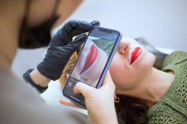 Makyaj sanatçısı akıllı telefon ekranında yaptığı çalışmaların fotoğrafını çekiyor, kadın dudaklarına kırmızı ve beyaz hat, kozmetikçi konsepti için sosyal medya profili uyguluyor