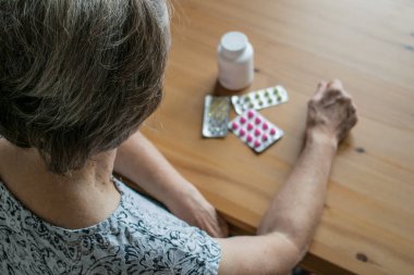 Hasta, yaşlı bir kadın evde, masanın yanına oturur ve haplara bakar.