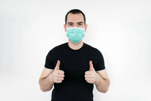 Erkek, genç, siyah tişört beyaz arka plan maskesi takıyor. Baş parmak hareketi. İki elinle koronavirüs sembolünü koru.