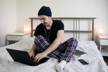  Ana konu odak dışı ya da net değil, genç erkek yatakta oturuyor ev içeriği yaratıcısı dizüstü bilgisayara konuşma kulaklıkları yazıyor.
