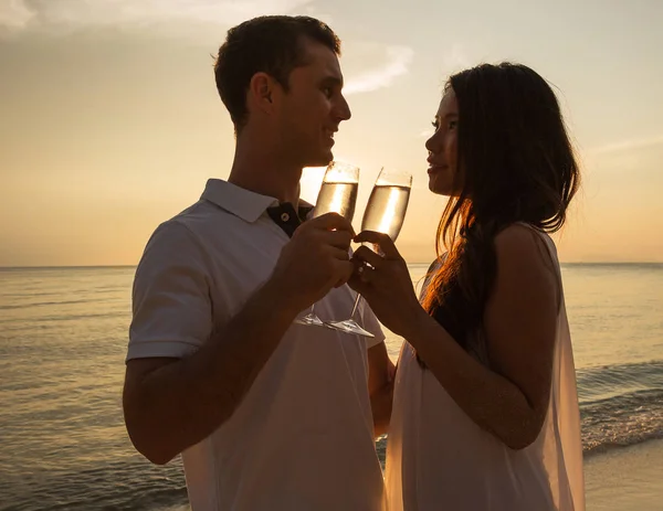 Romantic portrait of happy couple in love on the beach at the sunset on  tropical island. Looking each other. Smiling and holding in their hands glasses of champagne. Against the backdrop of the sea