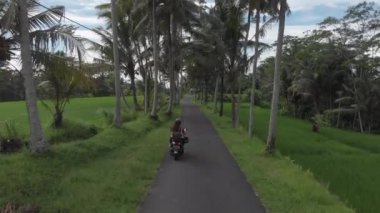 Bir adam ve bir kız palmiye ağaçlarının arasında motosiklet sürüyorlar.