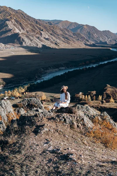 La chica en el sombrero con una mochila y un portátil se sienta en las piedras — Foto de Stock