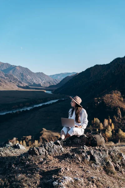 La chica en el sombrero con una mochila y un portátil se sienta en las piedras — Foto de Stock