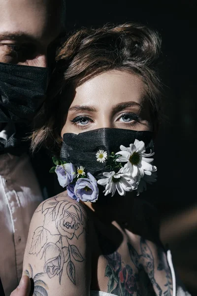 Gelin ve damat, siyah tıbbi maskeler takmış siyah arka planda taze çiçeklerle süslenmiş, spot ışıklarından ışınlar saçan gelinlik ve damatlar.