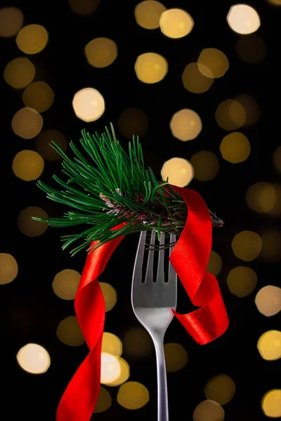 Creative Christmas or New Year card. Holidays or restaurant special dinner menu concept. Fork with fir branch and red satin ribbon with natural bokeh lights background