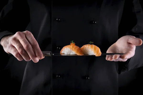 Sushi served on japanese knife in chef hands on dark background. Decorated salmon sashimi nigiri. Traditional japanese food. Copy space for text