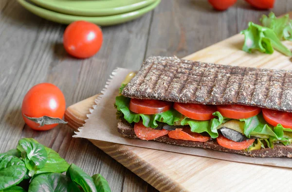 Panini - sandwich with brown bread, tomatoes, letuce and sausages on wooden background.