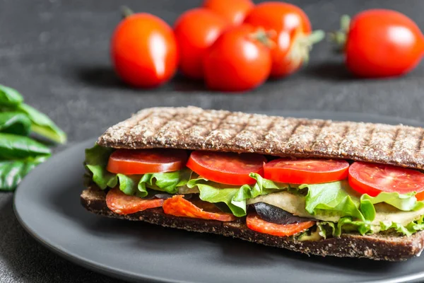 Panini - sandwich with brown bread, tomatoes, letuce, sausages, pieces of mozzarella and cup of coffee on black background. Front view