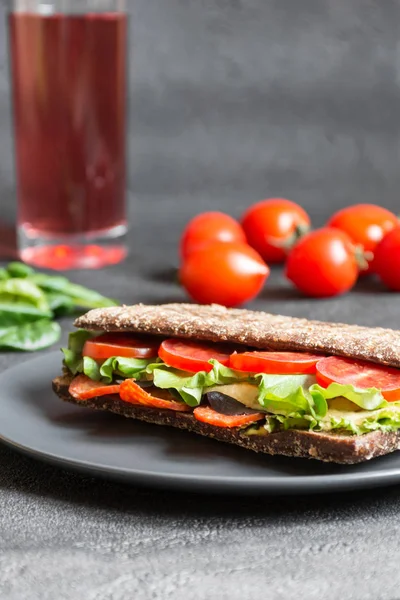 Panini - sandwich with brown bread, tomatoes, letuce, sausages, pieces of mozzarella and glass of juice on black background. Front view