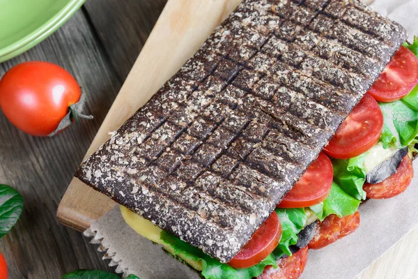 Panini - sandwich with brown bread, tomatoes, lettuce, sausages, pieces of mozzarella and cup of coffee on black background. Top view