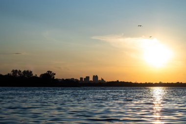 Şehrin arka planına karşı nehir kıyısında gün batımı.