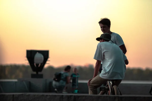 İki genç adam, biri bisikletin üzerinde oturuyor ikincisi ise bisikletin üzerinde gösteri yapmayı tartışmak için yanında duruyor. Gün batımının ve nehrin arka planında bir şehir parkında