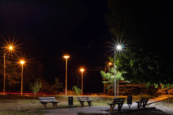 Empty benches in the evening park by the light of street lamps.