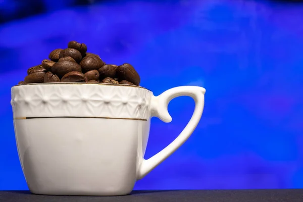 Kahve çekirdekleri ile doldurulmuş beyaz porselen bardak. Bulanık renk arkaplanı olan yakın plan..