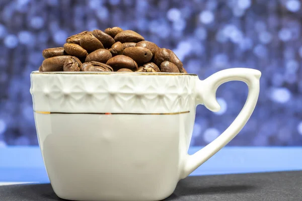 Kahve çekirdekleri ile doldurulmuş beyaz porselen bardak. Bulanık renk arkaplanı olan yakın plan..