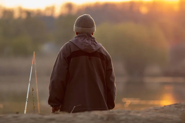 Şafak sökerken elinde terk edilmiş bir oltayla kıyıda duran şapkalı bir balıkçı. Bulanık arkaplan.