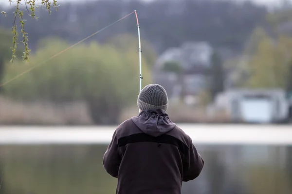 Nehrin arka planında elinde oltayla kıyıda duran şapkalı ve ceketli bir balıkçı. Bulanık arkaplan.