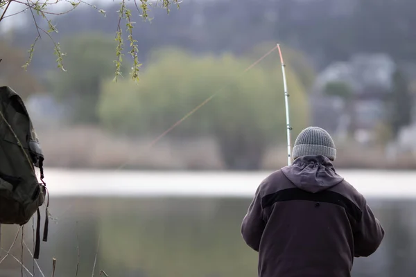 Nehrin arka planında elinde oltayla kıyıda duran şapkalı ve ceketli bir balıkçı. Bulanık arkaplan.