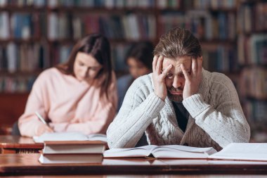 Sakallı adam kütüphanede kitap okurken yorgun görünüyor.