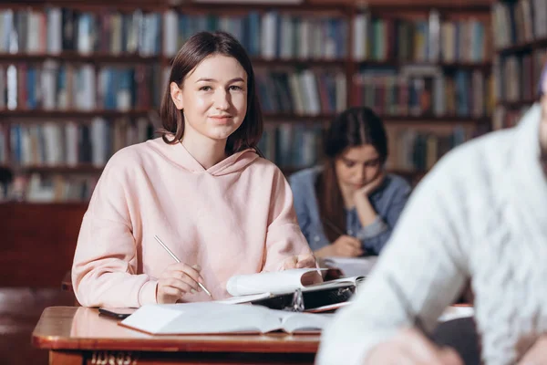 快乐的女孩坐在图书馆，看着相机 — 图库照片