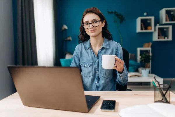 Evde dizüstü bilgisayarı ve sıcak kahvesiyle oturan genç bir kadın. — Stok fotoğraf
