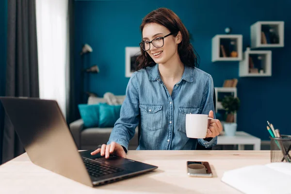 Neşeli kız dizüstü bilgisayarda çalışıyor ve kahve içiyor. — Stok fotoğraf