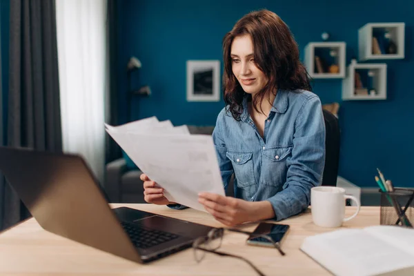 Щаслива дівчина сидить за столом з ноутбуком і робить паперові роботи — стокове фото