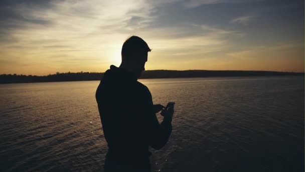 Mannen med smartphone vid solnedgången på floden bakgrunden. — Stockvideo