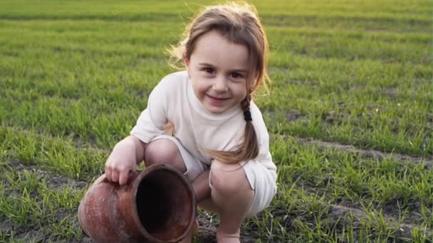 Simple country girl watering green grass with a clay pot on a background sunset. — Stock Video
