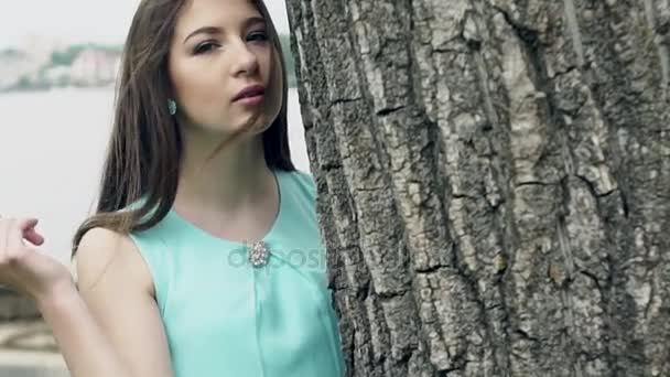 En blyg vacker flicka med vackra hår poserar på trädet mot bakgrund av staden. — Stockvideo
