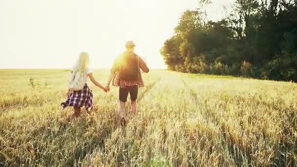 Dwoje kochanków podróży przez pole, trzymając się za ręce na tle wschód słońca na dzień jasny lato. — Wideo stockowe