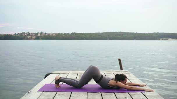 A beautiful brunette perform yoga exercises on the waterfront of the lake. — Stock Video