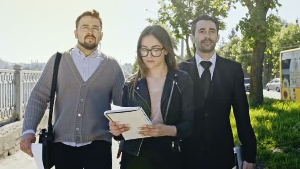 Tres profesionales, dos hombres y una mujer, caminando hacia la cámara en un camino junto a un camino con árboles, la mujer está leyendo notas . — Vídeos de Stock