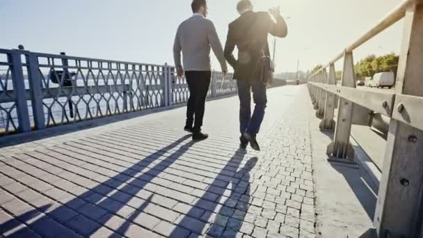 Dos hombres de negocios caminando por un puente lejos de la cámara con un zoom lento hasta que la cámara esté a la altura de ellos . — Vídeos de Stock