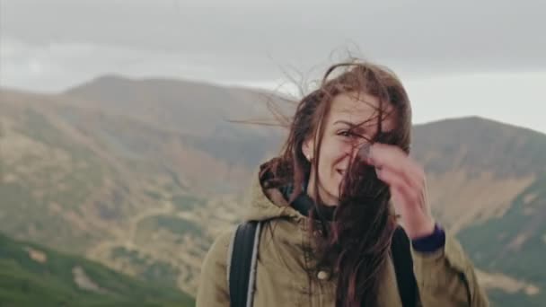 Attraente donna in piedi su una vetta di montagna con i suoi lunghi capelli che soffiano nel vento sorridendo e cercando di tenere i capelli fuori dal suo viso con la mano — Video Stock