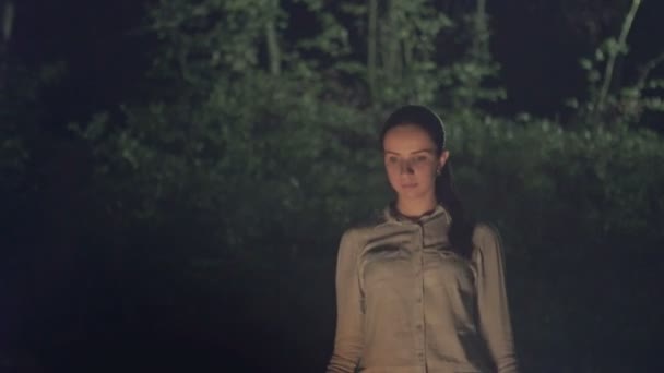 Девушка у костра в ночном лесу. На лице девушки сияет свет — стоковое видео