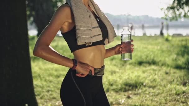 满足口渴。年轻愉快的妇女在体育锻炼以后休息在公园拿着瓶水和灰色毛巾 — 图库视频影像
