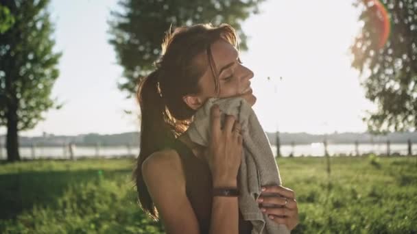 Menina fitness feliz limpa o rosto com uma toalha após exercícios pesados no pacote verde de verão no fundo do pôr do sol — Vídeo de Stock