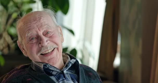 Портрет пожилого человека, сидящего на стуле и счастливо улыбающегося в камеру дома . — стоковое видео