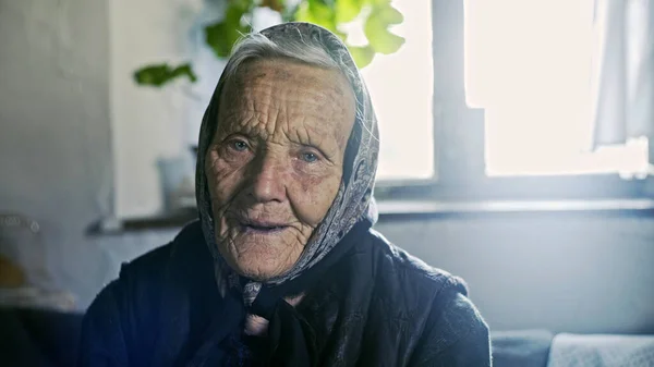 Amablemente anciana con una dulce sonrisa usando un pañuelo o pañuelo en la cabeza y un abrigo caliente en el interior de su casa con destello de sol desde una ventana de alta llave detrás de ella . — Foto de Stock