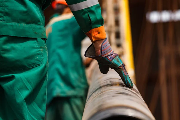 Oil workers in green and orange work wear and suede gloves on oil deposit with a rusted drilling pipes at summer day. Oil drilling rig. Close-up of worker hand. Second worker blurred on background.