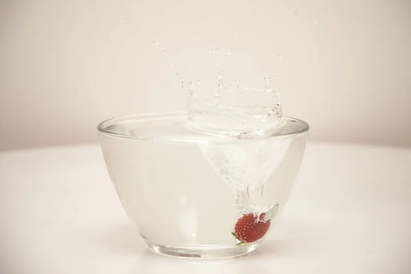 A single fresh strawberry is dropped into glass bowl filled in water on white background