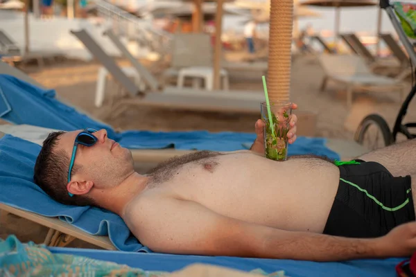 若い男は飲み物と一緒にラウンジのビーチに横たわっています — ストック写真