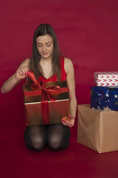 Mulher em um vestido vermelho abre um presente, muitos presentes ao lado dela — Fotografia de Stock